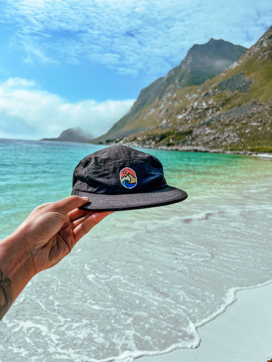 5 Panel Cap Classic – hånd holder den svarte capsen over turkis sjø