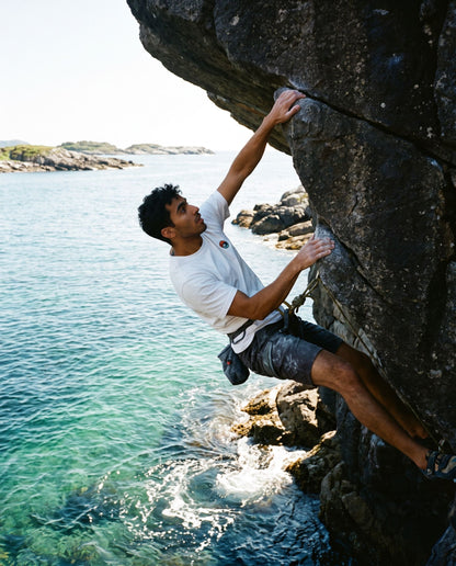 Let the Adventure Begin White Tee – mann klatrer uten tau over turkis sjø på kysten