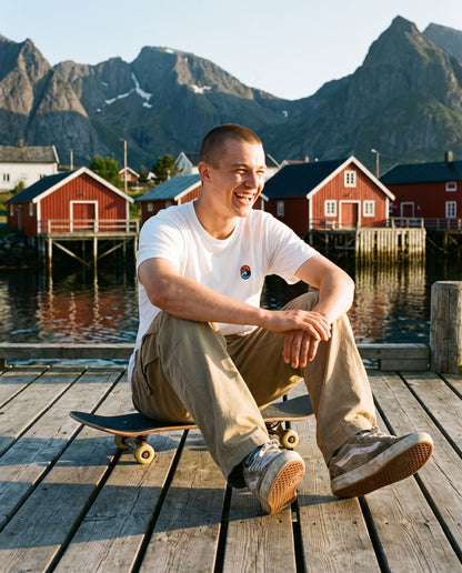 Let the Adventure Begin White Tee – smilende mann med skateboard på brygga i kveldssol i Lofoten