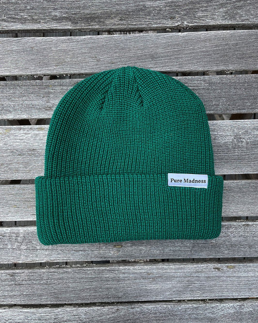 Green Captain Merino Beanie – grønn merinoull-lue med Pure Madness-merke på grå treplanker