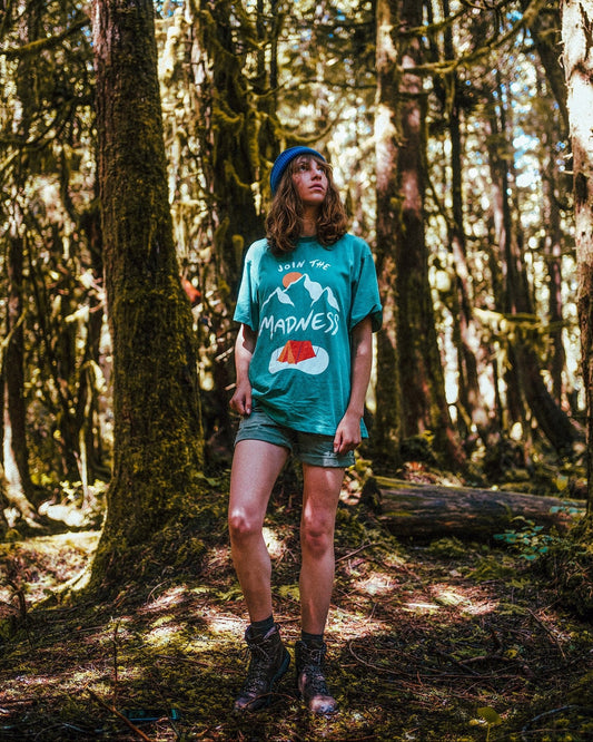 Join the Madness Teal Tee – kvinne i turkis t-skjorte og shorts i en mosegrodd skog