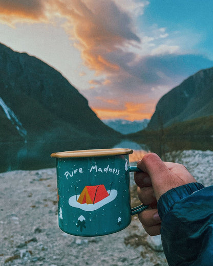 Winter Explorer Enamel Mug – hånd holder grønt emaljekrus med teltmotiv foran fjell i solnedgang